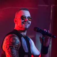 A male singer with a mohawk hairstyle and sunglasses performs on stage with his band. Wearing a sleeveless shirt revealing a large, intricate tattoo on his arm, he holds a microphone in one hand while making a gesturing pose with the other. The background is lit with red and purple stage lighting, perfectly capturing the energy of Sabaton's music.