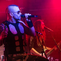 A man with a mohawk hairstyle and sunglasses sings into a microphone on stage, embodying the metal essence of Sabaton. He wears a black vest with metal plates and camo pants. Another man with long hair plays an electric guitar in the background. The stage is illuminated with red lights, creating a vibrant atmosphere for the band.