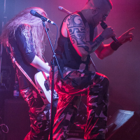 Two musicians from the heavy metal band Sabaton perform energetically on stage under red and purple lights. The vocalist, wearing sunglasses and a black tank top with extensive tattoos, sings passionately into a microphone. The guitarist, with long hair and clad in a black shirt, focuses on playing his white guitar. Both wear camouflage pants.