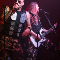 Two musicians performing on stage channel a Sabaton vibe. The lead singer, in sunglasses and a sleeveless vest with metallic patches, grips the microphone close to his mouth. Both he and the long-haired guitarist sport camouflage pants; the latter shreds on a white electric guitar against a dimly lit background.