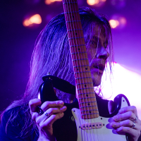 A guitarist with long hair, illuminated in purple light, holds an electric guitar vertically. The guitar has a white body and multiple strings. The background includes blurred round lights, enhancing the stage ambiance reminiscent of a Sabaton heavy metal band. The musician has a focused expression and wears rings and a black shirt.