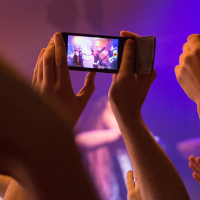 A person captures a Sabaton concert scene on a smartphone amongst a crowd of raised hands. The phone screen shows an image of the performer on stage under purple lighting. The atmosphere is lively, and the audience appears engaged, with arms raised in excitement.