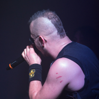 A person with a mohawk hairstyle is singing into a microphone on stage, exuding the heavy metal energy of Sabaton. They are wearing sunglasses and a black sleeveless shirt. A black wristband with a yellow emblem is on their left wrist, and three red marks are visible on their left shoulder. The background is dark and blurred.