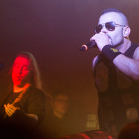 A male singer in sunglasses and sleeveless black attire is energetically performing on stage, holding a microphone close to his mouth. Channeling the intensity of Sabaton, the heavy metal band creates an electrifying atmosphere with vibrant stage lights and a foggy haze, while a guitarist, lit with red stage lighting, plays passionately in the background.