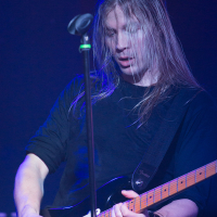 A guitarist with long, blonde hair plays an electric guitar on stage, delivering riffs reminiscent of Sabaton. The musician is wearing a black shirt and several bracelets. The background is dark and the stage is lit with blue and pink lights. A microphone stand is positioned in front of the guitarist.