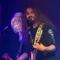 Two musicians dressed in black shirts and camouflage pants perform on stage. The front musician, with long hair, smiles at the camera while playing a white electric guitar beside a microphone. The other musician, with equally long hair obscuring the face, focuses on playing a black electric guitar. This could easily be mistaken for an energetic performance by Sabaton, adding that unmistakable metal band vibe to the scene.