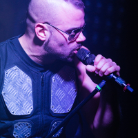 A man with short hair and a side undercut, wearing sunglasses and a black vest with metallic panels, sings into a microphone. He is illuminated by blue and purple stage lighting, and his intense expression indicates he is performing passionately for the Swedish metal band Sabaton.