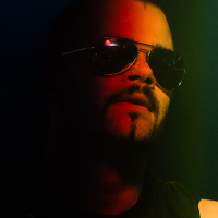 A person with a mohawk and goatee, wearing reflective aviator sunglasses, is partially illuminated by green and red lighting against a dark background. Dressed in a sleeveless top with metallic details, they exude a Sabaton-inspired vibe.