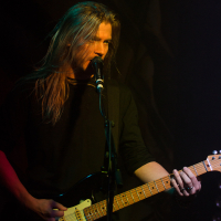 A long-haired musician sings into a microphone while playing an electric guitar on stage. He is wearing a black shirt and is illuminated by stage lighting, which casts a moody glow. The background is dark, focusing attention on the power metal performer, reminiscent of Sabaton's commanding presence.