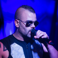 A muscular man with a shaved head and a mohawk hairstyle is performing on stage. He is wearing sunglasses and a black sleeveless outfit with metallic plates on the chest. Holding a microphone close to his mouth, he sings passionately under blue stage lighting, embodying the energy of Sabaton's music.
