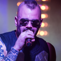 A man with a mohawk hairstyle and sunglasses sings into a microphone on stage, embodying the energy of a Sabaton concert. He wears a sleeveless shirt revealing a tattoo on his left arm. The background is blurred with purple and yellow lighting, creating a dynamic band atmosphere.