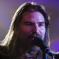 A person with long brown hair and a beard is singing into a microphone on stage, reminiscent of Sabaton's energetic performances. The dramatic lighting, with a purple hue in the background, creates an intense atmosphere. Wearing a necklace and a black shirt, they appear focused and passionate during their performance.