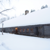 A long wooden cabin with a snow-covered roof stands in the snowy landscape of Winter Solstice 2010. Warm light glows through its windows, contrasting with the cold, white surroundings. A wooden door is on the right side, and a snow-covered tree is seen to the left. A lamppost is near the entrance of the cabin.