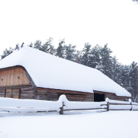 A wooden cabin with a steep, snow-covered roof stands amidst a snowy landscape during the Winter Solstice in 2010. The surrounding area, including a wooden fence in the foreground, is blanketed in thick snow. Tall evergreen trees with snow-laden branches frame the scene, creating a secluded winter atmosphere.