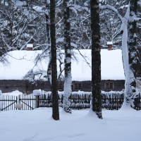 A snow-covered wooden house is nestled in a forest. The house, dating back to 2010, has a white, snow-laden roof and brown walls. Bare trees and a rustic wooden fence with snow drifts surround the building. The ground is blanketed in a thick layer of snow, creating a serene Winter Solstice scene.