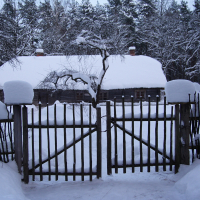A rustic wooden fence gate stands open, leading to a snow-covered yard with a thatched-roof cottage in the background. The roof and surroundings are heavily blanketed in snow, including the tree branches and shrubs. Tall pine trees in the background add depth to the winter solstice scene.