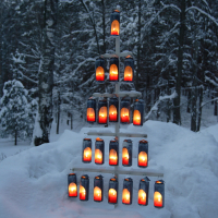 A tiered pyramid of lit lanterns stands on a snow-covered ground amidst a forest of snow-laden trees. The glowing lanterns create a warm, golden light contrasting sharply with the cold, blue-tinted snow and dark tree trunks, giving the scene a serene and festive atmosphere reminiscent of Winter Solstice 2010.