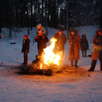 A group of people, bundled in winter clothing, stand around a large bonfire in a snowy forest during the 2010 Winter Solstice. One child is seated on an adult's shoulders. Skis are visible on one person to the left. The scene is illuminated by the firelight, contrasting with the dark, snow-covered trees in the background.
