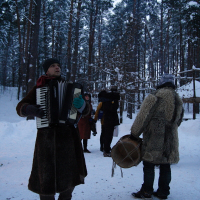 A group of people stands in a snowy forest, celebrating the Winter Solstice. The central figure, wearing a long coat, plays an accordion, while another in a fur coat holds a drum. Other individuals in the background are dressed warmly and seem engaged in conversation. Snow-laden trees surround the scene.