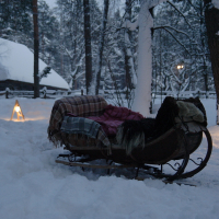 A wooden sleigh covered with blankets sits in a snowy forest. The area is softly illuminated by triangular lanterns on either side of the sleigh. Snow-covered trees and a rustic, snow-covered cabin are visible in the background, evoking memories of the serene Winter Solstice 2010.