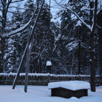 A snow-covered scene featuring a traditional wooden well with a long pole for drawing water, situated in a fenced woodland area during the 2010 Winter Solstice. Tall trees stand in the background, their branches dusted with snow. The sky is a soft, twilight blue, illuminating the serene winter landscape.