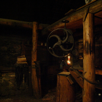 A dimly lit room with wooden beams and rough stone walls. In the foreground, an old-fashioned hand-operated grinding wheel is mounted on a wooden stand, its small candle flickering like it's waiting for the Winter Solstice. Stacks of wood and various tools are visible in the background. The floor is made of worn bricks.