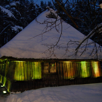 A rustic wooden cabin with a snow-covered steep thatched roof, illuminated by soft green and yellow lights at dusk. The cabin is surrounded by snow and tall, dark trees which create a serene Winter Solstice atmosphere. Thin branches in the foreground are also lightly dusted with snow.