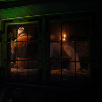 View through a darkened, frost-covered window with wooden trim, lit by a dim green light. Inside, on the night of the Winter Solstice in 2010, a figure in a brown coat and knit hat stands next to another figure in white, both facing away, with a warm light source illuminating the interior partially.
