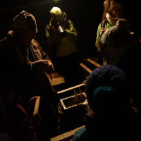 A group of people dressed warmly in hats and jackets are gathered indoors under a single overhead light on the 2010 Winter Solstice. One person is preparing or handling something at a wooden table, while another in bright clothing stands in the background, facing away and drinking from a thermos. The scene appears dimly lit.