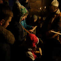 A group of people, including children and adults dressed in winter clothing, are gathered closely together indoors. They appear to be engaged in a Christmas or Winter Solstice celebration, with some holding wooden handmade crafts. The festive atmosphere is enhanced by candles in the background, reminiscent of a cozy 2010 gathering.