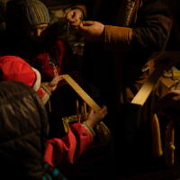In a dimly lit setting during the Winter Solstice of 2010, people gather around a table. A person in a brown coat demonstrates a craft to children wearing winter clothing, including a striped hat and red jacket. Various handmade candles are on the table, illuminated by nearby candles, creating a cozy atmosphere.