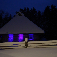 A snow-covered rustic cabin with a thatched roof is illuminated by purple lights at night, evoking memories of the tranquil Winter Solstice in 2010. A snow-laden wooden fence graces the foreground while tall trees are silhouetted in the background, enhancing the calm and serene wintry atmosphere.