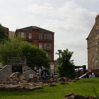 A group of tourists relax on stone steps in an urban park surrounded by greenery, with traditional red sandstone buildings under a cloudy sky in the background, in Scotland.