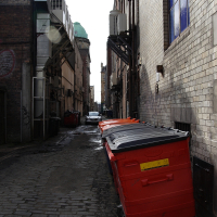 A narrow urban alleyway in Scotland lined with brick buildings and large red commercial waste bins under a partly cloudy sky, showcasing a gritty, utilitarian aspect of city infrastructure.