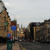 A bustling city street descends towards a historic skyline peppered with spires and domes under a cloudy sky; signs, vehicles, and pedestrians animate the scene, encapsulating urban life in Scottish culture