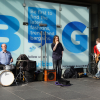 Street musicians perform outside a shopping center in Scotland; a drummer and two guitarists engage with passing pedestrians, their enthusiasm contrasted against the backdrop of commercial advertisements.