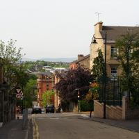 A descending urban street in Scotland, lined with historic buildings, featuring a blossoming tree and a view extending towards a green landscape under a hazy sky.