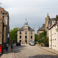 A cobblestone street leads towards an imposing stone building with a central clock tower, flanked by traditional architecture under a cloudy sky, while pedestrians and vehicles share the quaint urban setting, perfectly embodying