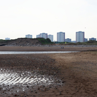 A serene beach with a rippled tidal pool in the foreground, a solitary figure walks along the shore, underlining Scotland's travel allure, with an urban skyline of high-rise buildings in the distance under