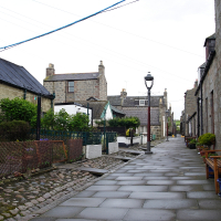 A quaint cobblestone alley flanked by traditional stone buildings and modest front gardens under an overcast sky, offering a glimpse into the charming architectural heritage of a historic area in Scotland.