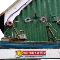 A wooden model of a boat is displayed atop a white ledge, adorned by a colorful sign proclaiming "smile, you're in Aberdeen - Scotland's happiest city," against a rustic green background.