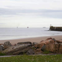 A serene beach scene in Scotland with scattered boulders in the foreground, calm tide gently washing ashore, and distant ships near a protective breakwater under a soft sky.