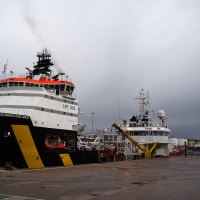 A supply vessel and a tugboat moored at a bustling industrial harbor in Scotland against a backdrop of overcast skies, with port infrastructure and shipping equipment visible in the surroundings.