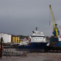 A moody, overcast sky hangs over a bustling seaport in Scotland, where a large blue ship docks near a towering yellow crane, with storage tanks and various maritime equipment scattered around the industrial