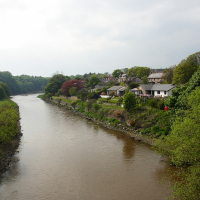 A serene riverside village in Scotland, cloaked in greenery, with houses nestled among the trees overlooking the tranquil waterway, under an overcast sky.