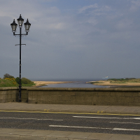 A solitary lamppost stands by a seaside road in Scotland, overlooking a tranquil beach, with a ship visible on the horizon under a vast, cloudy sky.
