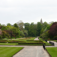 A serene, manicured park in Scotland with lush green lawns, vibrant flower beds, and diverse trees. A central path leads to an ornate structure in the distance, under a cloudy sky.