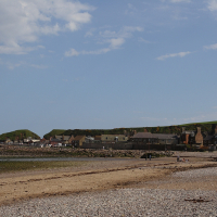 A tranquil beach at low tide with a quaint coastal village stretching across the background under a partly cloudy sky in Scotland, as visitors enjoy the shoreline.
