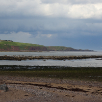 A tranquil seaside landscape under a dramatic sky, with exposed rocks at low tide, a sandy shore in the foreground, and verdant cliffs in the distance, evoking Scotland's rich history and captivating travel