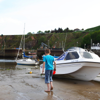 A boy walks along a muddy shoreline in Scotland toward a beached white-and-blue boat named "Lucky Lady," with other boats and a green hillside in the background, highlighting the beauty of travel